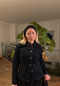 Dunkelblaue Jeansjacke mit tailliertem Schnitt, silbernen Knöpfen, Kragen und langen Ärmeln. Im Hintergrund sind grüne Pflanzen und ein Geländer zu sehen.