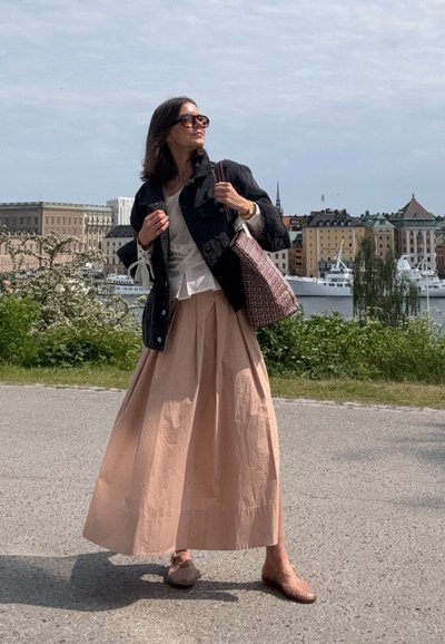 Larga falda beige con plisados, blusa blanca, chaqueta de mezclilla negra y bolsa de mano estampada. Entorno al aire libre, con edificios y agua de fondo.