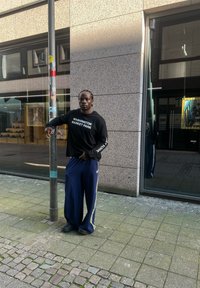 Μαύρο μακρυμάνικο πουκάμισο με κείμενο "WASHINGTON MARKET PARK", χαλαρές ναυτικές μπλε παντελόνες με λευκές ρίγες και μαύρα αθλητικά παπούτσια, στέκεται δίπλα σε μια κολώνα του δρόμου.