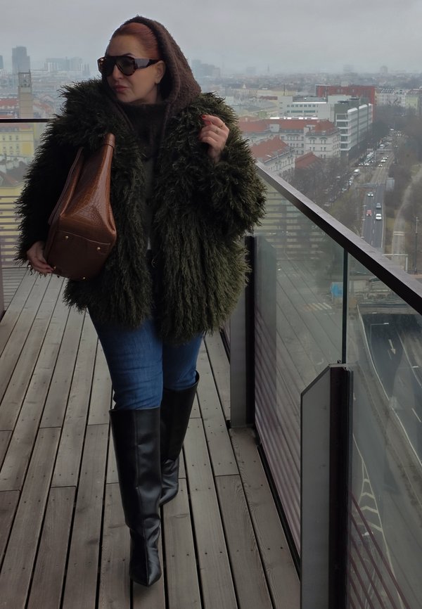 Kvinde iført mørkegrøn pelsjakke, brun pandebånd, solbriller og sorte støvler gående på tagterrasse med bylandskab i baggrunden.