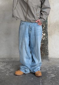 Ruda ir balta languota itin didelio dydžio marškiniai, šviesiai mėlyni platūs džinsai ir rudos darbo batai su tekstūruota padu, stovintys prie neutralaus fono.