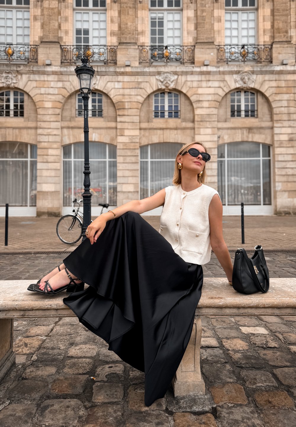 A woman sits on a stone bench, wearing a sleeveless cream top, a flowing black skirt, and strappy black sandals, with a black handbag nearby.