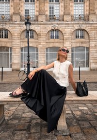 A woman sits on a stone bench, wearing a sleeveless cream top, a flowing black skirt, and strappy black sandals, with a black handbag nearby.