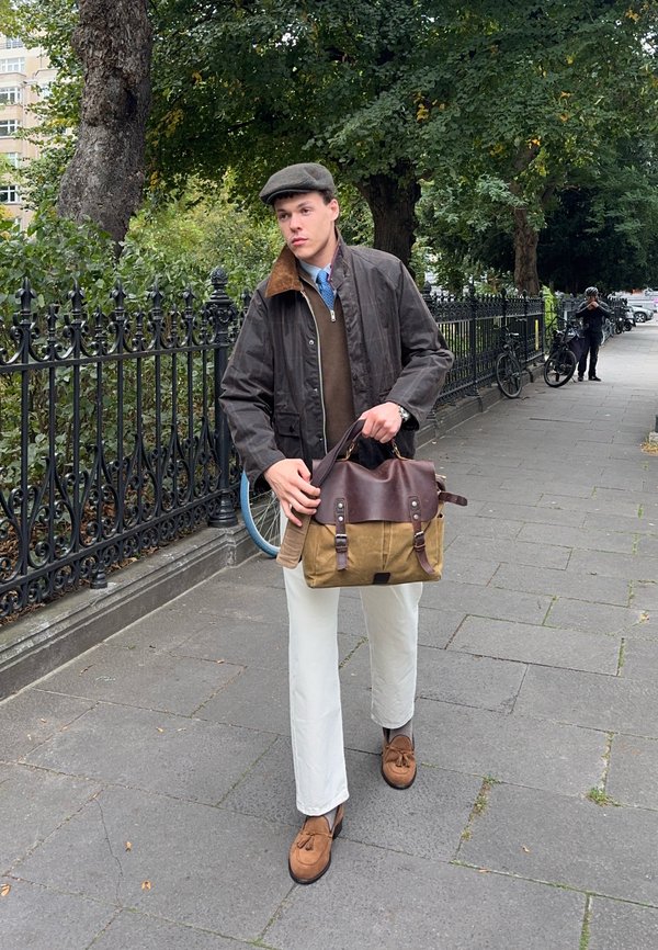Un homme portant une casquette plate et une veste brune tient un sac en cuir et en toile sur un trottoir urbain bordé d’arbres, près d’une grille de parc et de vélos.
