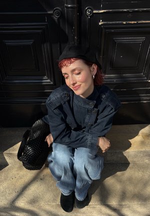 Joven con pelo corto y rojo, boina negra, chaqueta vaquera y vaqueros sentado con las piernas cruzadas en unos escalones a la luz del sol con un bolso negro tejido.