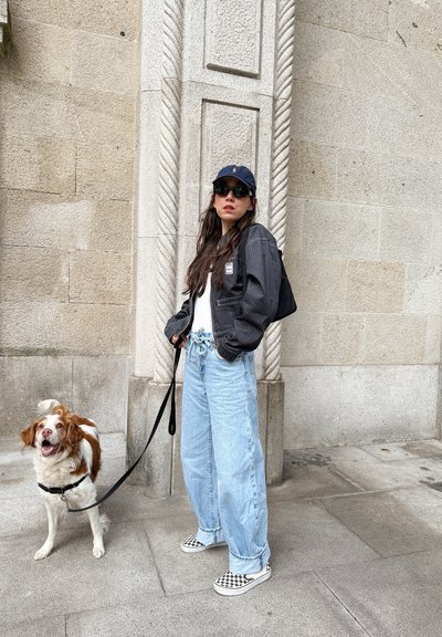 Persona vestida con una chaqueta a rayas negra, camisa blanca y jeans de mezclilla azul claro, combinada con zapatos tipo slip-on de cuadros, se encuentra al lado de un perro con correa.