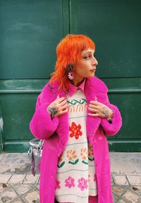 Abrigo rosa fucsia difuso, suéter crema con patrón de diseños florales en rojo y verde, y bolso plateado. Cabello naranja y pendientes florales.