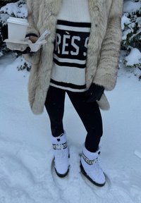 Personne debout dans la neige portant des bottes de neige blanches, des leggings noirs, un pull rayé, un manteau en fourrure et des gants noirs, tenant du café et des encas.