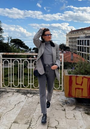 Blazer a cuadros grises con un cuello negro sobre un suéter de cuello alto gris, combinado con jeans grises desgastados y zapatos negros. Entorno al aire libre con vista.