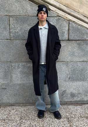 Joven con abrigo largo negro, suéter gris, vaqueros holgados, botas negras y un gorro negro con logo blanco, de pie contra una pared de piedra.