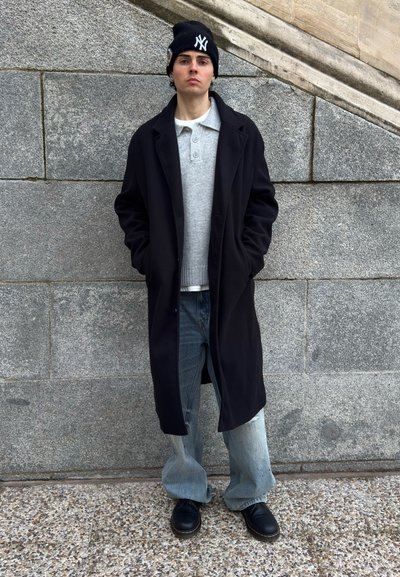 Joven con abrigo largo negro, suéter gris, vaqueros holgados, botas negras y un gorro negro con logo blanco, de pie contra una pared de piedra.