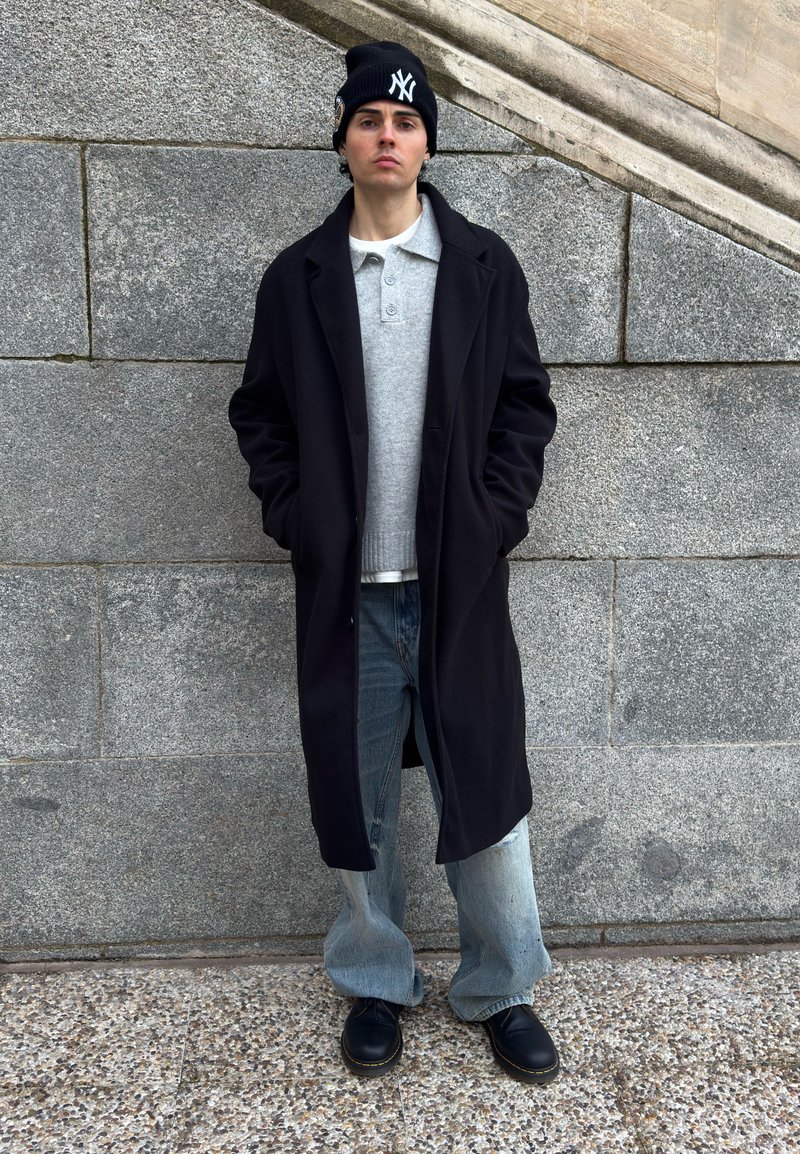 Jeune homme portant un long manteau noir, un pull gris, un jean ample, des bottes noires et un bonnet noir avec un logo blanc, debout contre un mur en pierre.