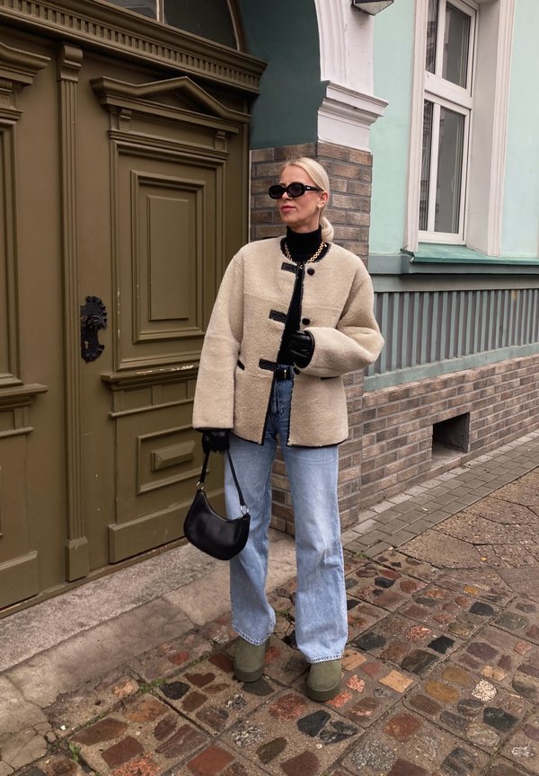 Beżowa polarowa kurtka z czarnymi guzikami, czarny golf, jasnoniebieskie dżinsy z szerokimi nogawkami, zielone buty na platformie i mała czarna torebka.