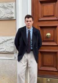 Jūrinės tamsiai mėlynos spalvos dvigubo užsegimo švarkas, dėvimas su šviesiai mėlyna marškiniais, tamsia raštuota kaklaraiščiu ir smėlio spalvos kelnėmis. Stovint šalia medinių durų su bronziniais furnitūromis.