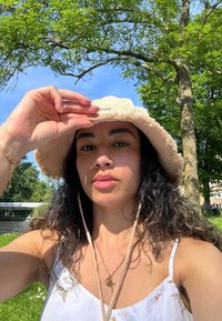 Beige bucket hat with frayed edges and chin straps, worn with a white dress featuring floral embroidery. The background includes lush green foliage and a blue sky.