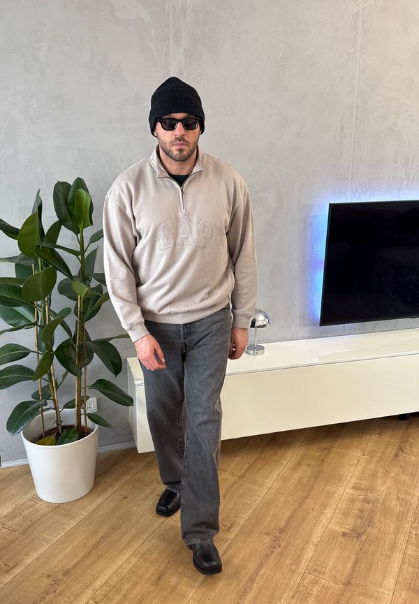 Mees, kes kannab musta beanie't, päikeseprille, beeži GAP pusa, halli teksapükse ja musti kingi, kõnnib siseruumides taime ja seinale paigaldatud teleri lähedal.