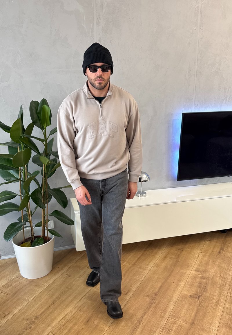 Homme portant un bonnet noir, des lunettes de soleil, un sweat-shirt beige GAP, un jean gris et des chaussures noires, marchant à l'intérieur près d'une plante et d'une télévision murale.