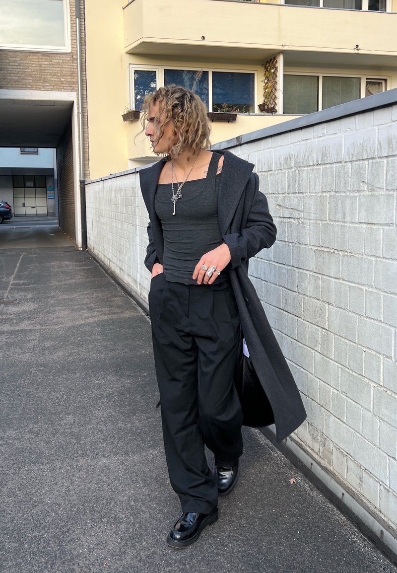 Black overcoat worn over a fitted grey tank top, paired with loose black trousers and shiny black shoes. Distinctive silver jewellery highlights.