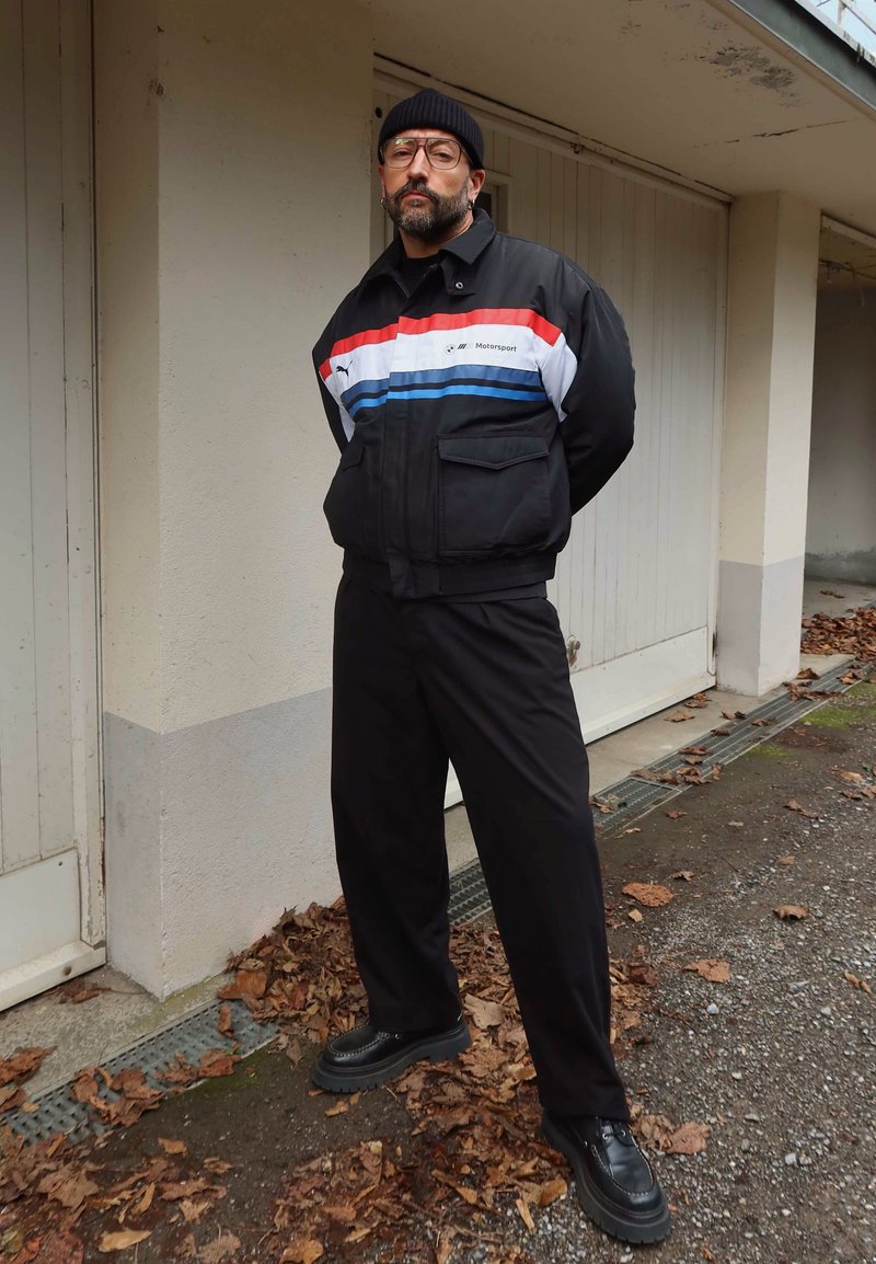 Homme portant un bonnet noir, des lunettes et une veste noire avec des bandes rouges, blanches et bleues, debout les mains dans le dos près des portes du garage.