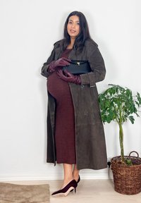 Femme enceinte en robe marron et long manteau gris tenant une pochette noire, portant des gants violets et des talons assortis, debout à côté d'une plante en pot.