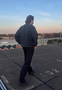 Μαύρο oversized μπουφάν, μαύρο παντελόνι και μαύρα παπούτσια. Υπαίθρια σκηνή στη στέγη με σκυρόδεμα και μια κιγκλίδωμα στο φόντο.