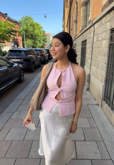 Top halter rosa con detalles recortados y acentos de lazo, combinado con una falda blanca fluida y un bolso de mimbre. Escenario en la acera.