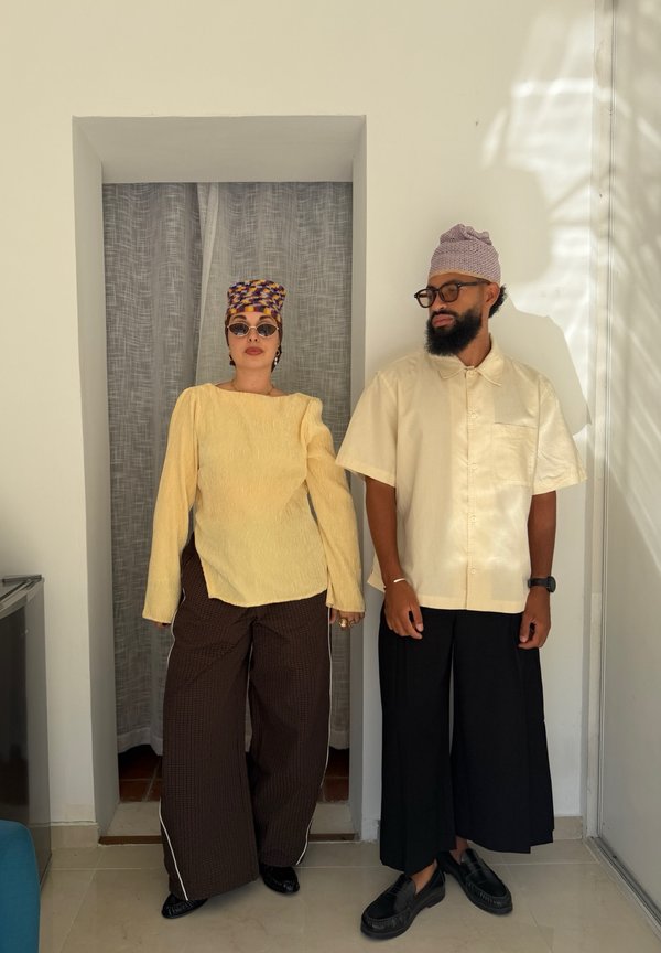 Deux personnes debout à l'intérieur portant des vêtements amples et stylés ainsi que des foulards à motifs, avec des lunettes et des hauts de couleur neutre.