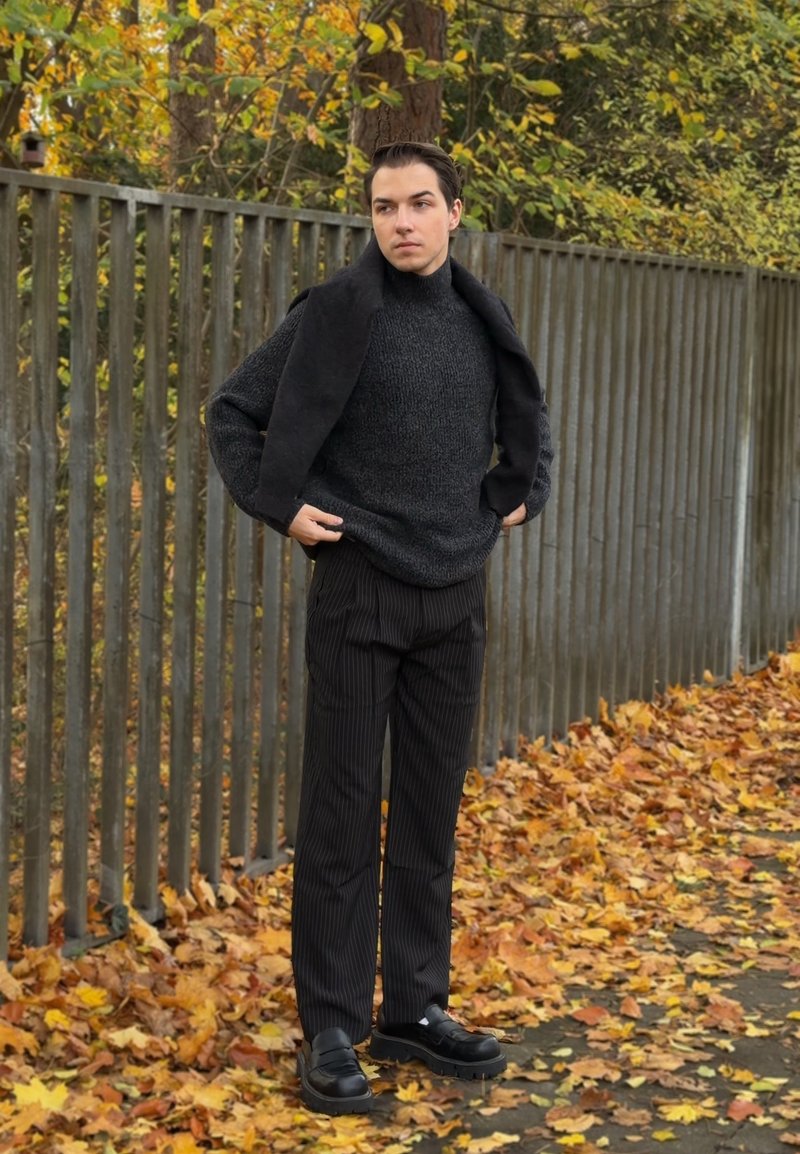 Maglione nero testurizzato indossato sotto un cappotto scuro, abbinato a pantaloni neri a righe sottili e scarpe nere robuste, sullo sfondo di un contesto autunnale con foglie.