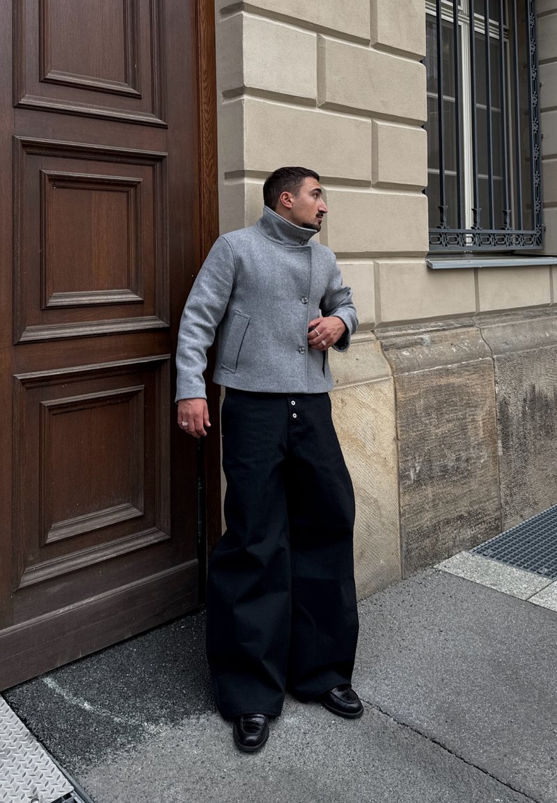 Homme portant une veste grise et un pantalon large noir se tient près d'une porte en bois et d'un mur en pierre avec une fenêtre à barreaux métalliques dans une rue de la ville.