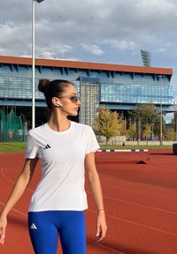 Ung kvinne som har på solbriller og treningsklær, står på en rød løpebane med en stadionbygning og trær i bakgrunnen.