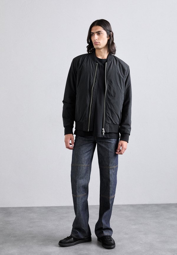 Giacca bomber nera con chiusura a zip, maniche lunghe e polsini a coste, abbinata a jeans denim scuri con cuciture a vista. Sneakers nere.