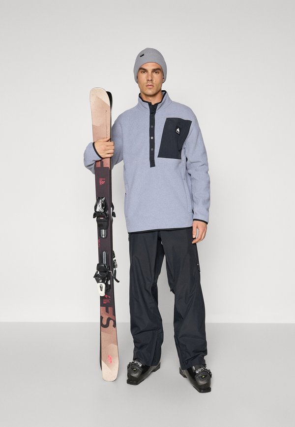 Fleece genser i lyseblå med en svart brystlomme, svarte skibukser og en grå lue. Holder brune og svarte ski med bindinger.