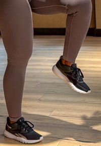 Piernas con leggings malva y zapatillas negras y blancas, un pie en el suelo y el otro levantado, de pie sobre un suelo de madera en interiores.