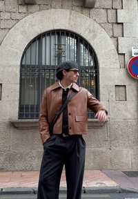 Casaco curto de couro castanho com gola, dois botões frontais, usado sobre um suéter preto, combinado com calças escuras e uma bolsa transversal.