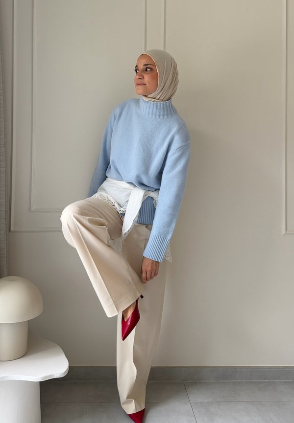 Une femme portant un hijab beige, un pull bleu, un pantalon crème et des chaussures rouges à talons pose devant un mur clair, un pied levé à côté d’une table blanche.