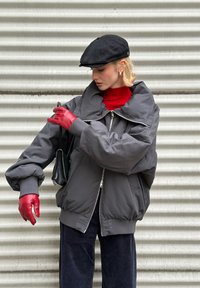 Giacca bomber grigia oversize con collo alto, cerniera argento, abbinata a dolcevita rossa e guanti in pelle rossa. Cappello piatto nero e borsa.