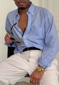 Chemise bleu clair à manches longues, partiellement déboutonnée, avec une poche. Pantalon beige clair. Personne tenant des lunettes de soleil et portant une montre en or.