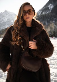 Γυναίκα με μακριά κυματιστά μαλλιά που φοράει μεγάλα γυαλιά ηλίου, σκούρο καφέ γούνινο παλτό και κρατάει καφέ δερμάτινη τσάντα που ταιριάζει, σε χιονισμένα βουνά.