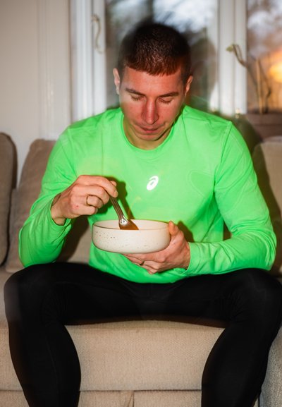 Hombre con camiseta de manga larga verde sentado en un sofá, sosteniendo un tazón y revolviendo comida con una cuchara, mirando hacia abajo al tazón.
