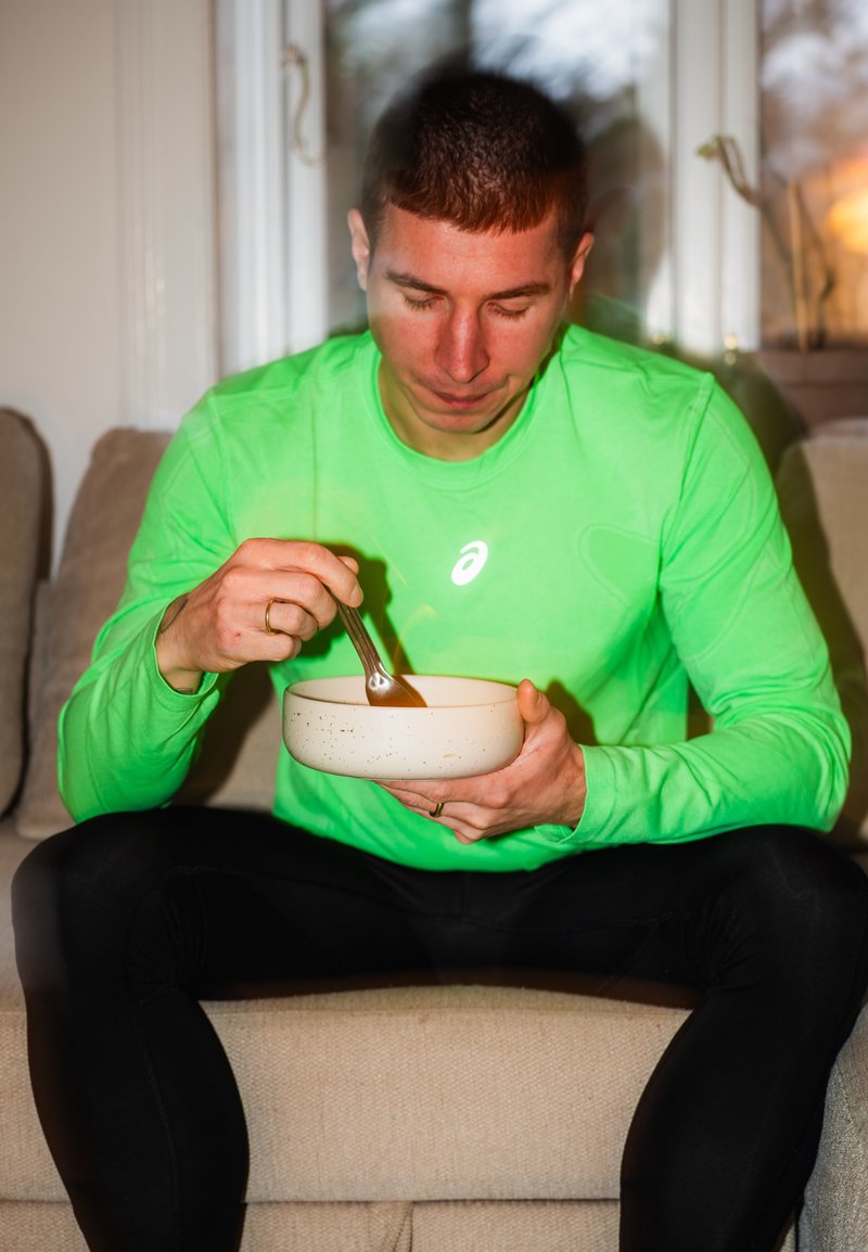 Άνδρας με πράσινο πουκάμισο μακρυμάνικο κάθεται σε έναν καναπέ, κρατώντας ένα μπολ και ανακατεύοντας φαγητό με ένα κουτάλι, κοιτάζοντας προς τα κάτω το μπολ.