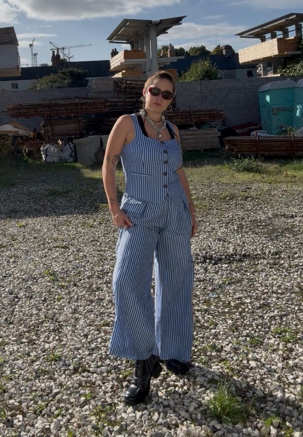 Una mujer con gafas de sol y un mono de rayas azules y blancas está de pie sobre grava en una obra.