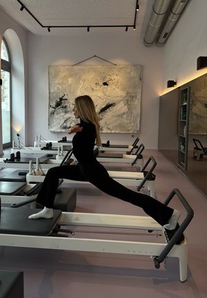 Reformer Pilates berendezés fehér vázzal és fekete párnázással. Egy személy bemutatja a kilépés pozíciót a reformeren. Világos rózsaszín padló.