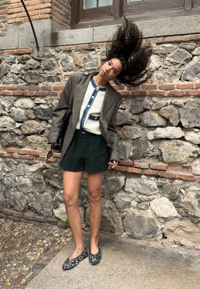 Mujer con cabello largo y rizado se echa el pelo hacia atrás, lleva blazer gris, camisa blanca abotonada, pantalones cortos negros y zapatos planos estampados, de pie junto a un muro de piedra.