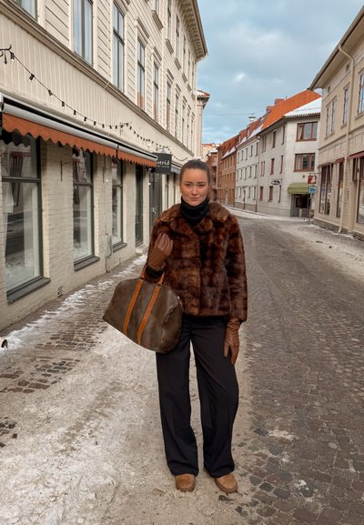 Mujer con chaqueta de piel marrón, guantes y pantalones negros, de pie en una calle adoquinada cubierta de nieve, bordeada de edificios bajo un cielo nublado.