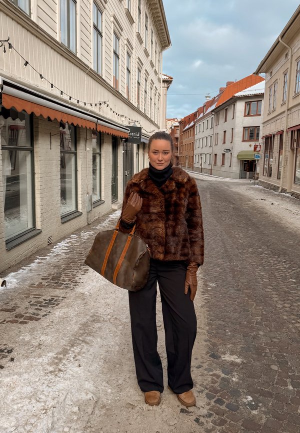 Femme portant une veste en fourrure marron, des gants et un pantalon noir, debout dans une rue pavée enneigée bordée de bâtiments sous un ciel nuageux.