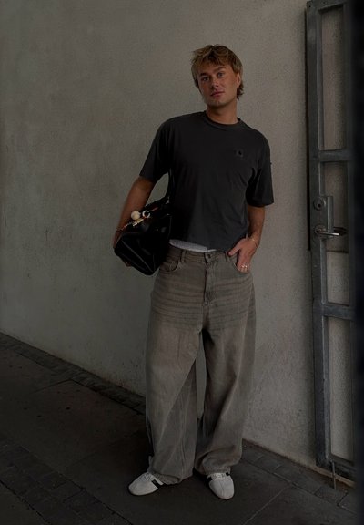 Camiseta gris cortada, pantalones de mezclilla beige sueltos y zapatillas blancas. Bolso negro con cuentas decorativas. De pie contra una pared texturizada.