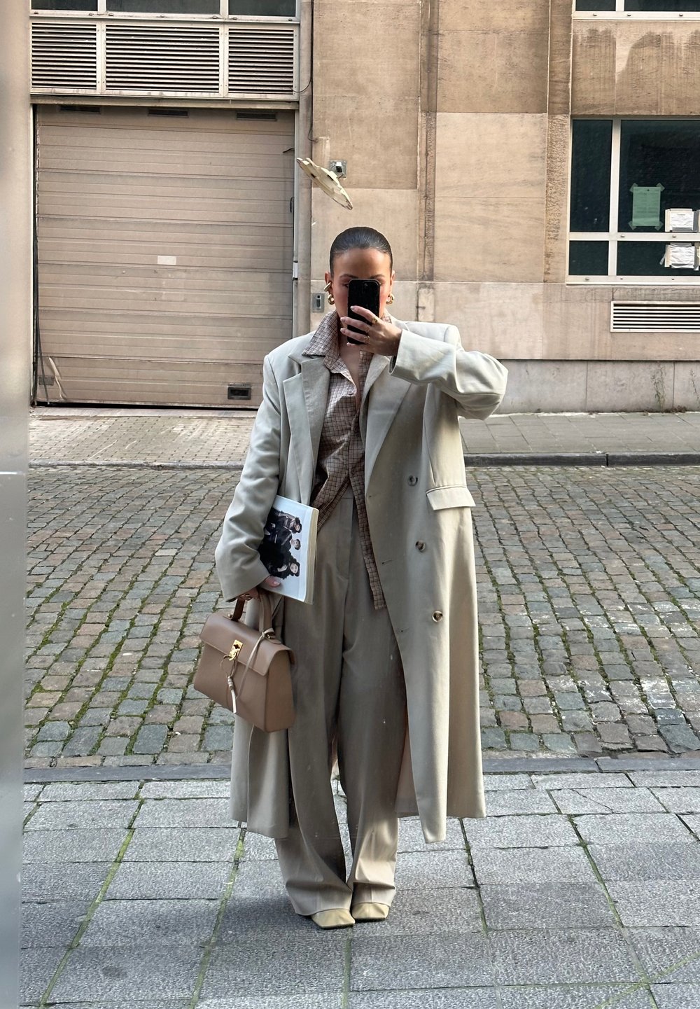Beige trenchcoat met grote knopen, een geruit shirt eronder, wijde broek en een lichtbruine handtas. Houdt een tijdschrift voor een spiegel.