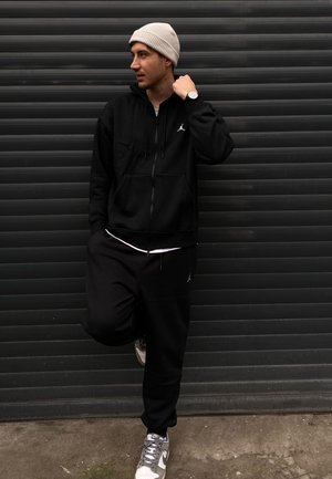 Sudadera con zipper negra y joggers con el logo de Jordan, combinados con un gorro ligero gris y una camiseta blanca, completados con zapatillas gris y blancas.