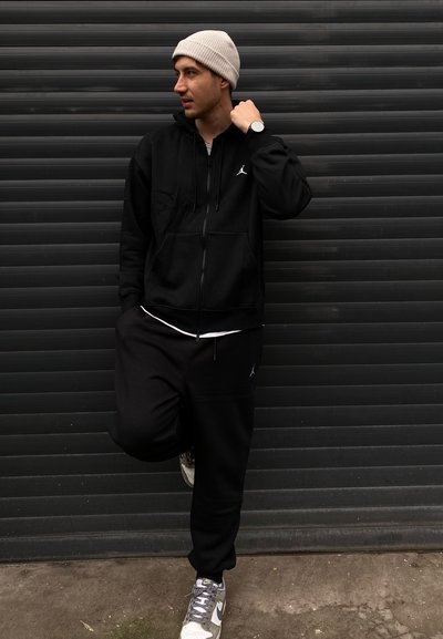 Sudadera con zipper negra y joggers con el logo de Jordan, combinados con un gorro ligero gris y una camiseta blanca, completados con zapatillas gris y blancas.