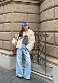 Młoda kobieta w puszystej kurtce, niebieskiej czapce, szerokich dżinsach i czarnych butach, trzymająca zielony napój, opierająca się o ozdobne brązowe ogrodzenie przy kamiennym murze.